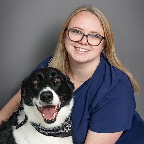 A smiling woman with long blonde hair and glasses, wearing a navy blue top, poses with a happy black and white dog wearing a patterned bandana in front of a plain gray background.