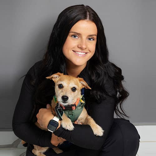 A woman with long dark hair, wearing a black outfit and a smartwatch, smiles while hugging a small brown dog in a green harness against a plain gray background.