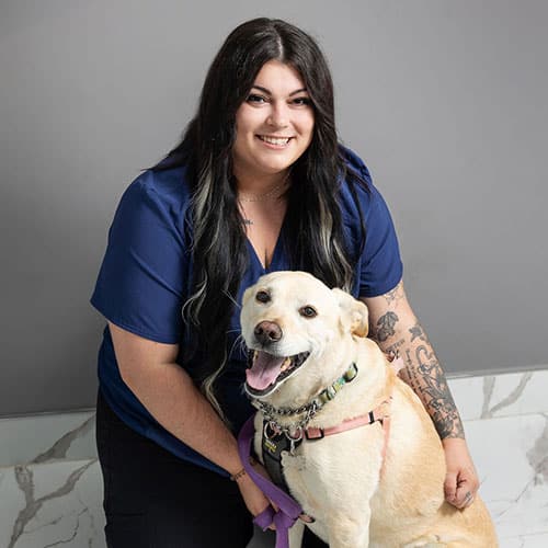 A smiling woman with long dark hair and tattoos, wearing a blue top, poses with a happy yellow Labrador Retriever wearing a pink harness and leash, against a light gray background.