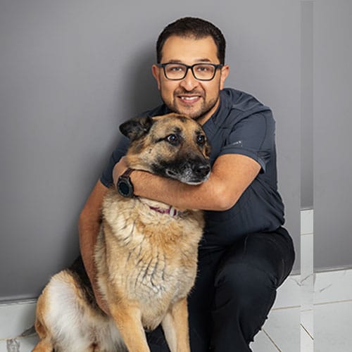 A man wearing glasses and a dark shirt is kneeling and hugging a large German Shepherd dog. They are both posed in front of a plain gray wall and are looking at the camera, appearing happy and relaxed.
