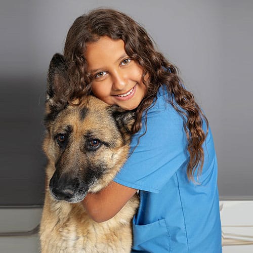 A smiling girl with long curly hair wearing blue scrubs hugs a large German Shepherd dog, both looking at the camera against a plain gray background.