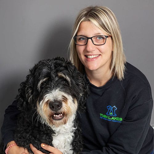 A smiling woman wearing glasses and a "Twin Lakes Veterinary Hospital" sweatshirt sits next to a large, fluffy black and white dog. Both are looking at the camera against a plain gray background.