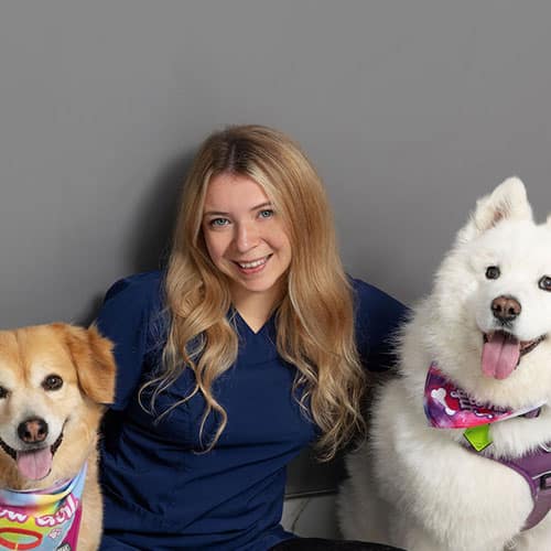 A smiling woman with long blonde hair, wearing navy blue scrubs, sits between two happy dogs wearing colorful bandanas, posing in front of a plain gray background.